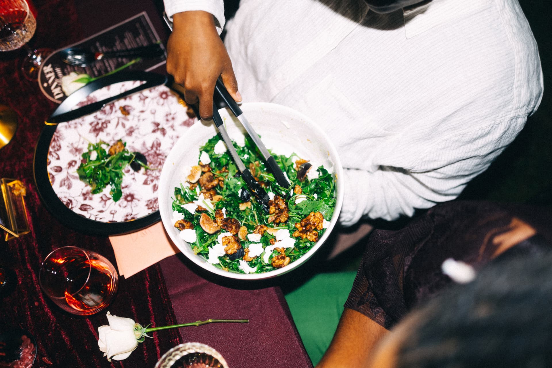 A fresh arugula salad with figs, walnuts, and soft cheese being served from a large communal bowl onto a patterned plate at a burgundy-draped table