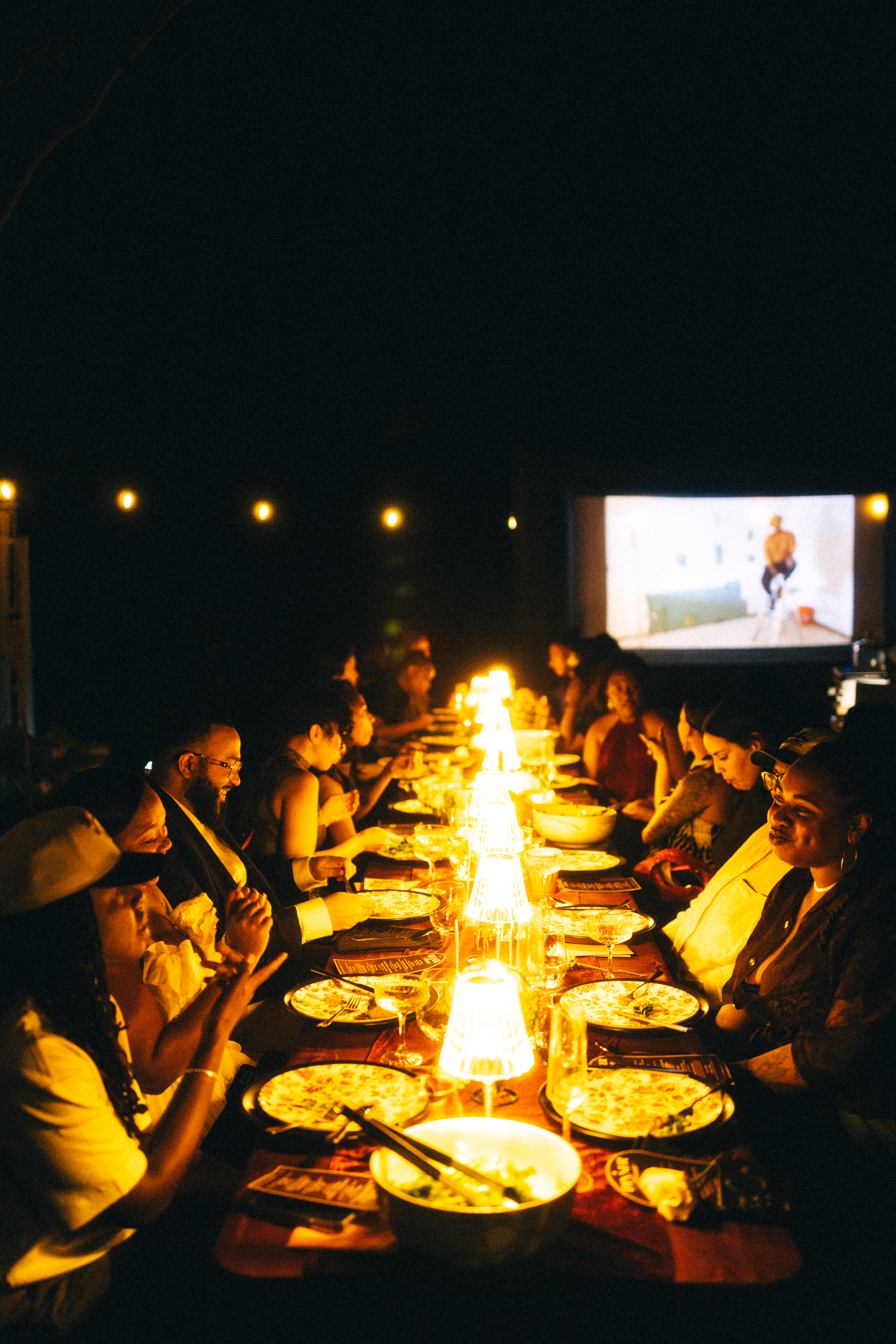The long dinner table lit by lamps, with a film projection glowing at the far end